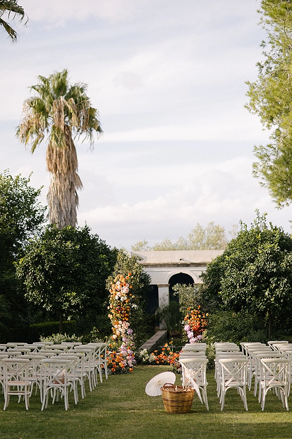 Outdoor ceremony setup with garden wedding ceremony aisle lined by white chairs, floral pillars, and scattered petals on a lawn near white arches