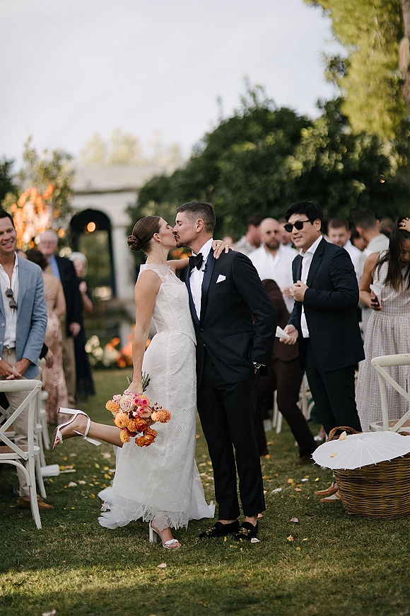 Wedding kiss portrait of bride and groom kissing in the aisle, bride holding a colorful bouquet as guests cheer in a garden setting.