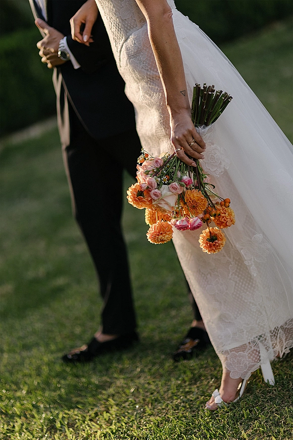 Bride and groom portrait with wedding bouquet close up, showcasing lace dress and rings as they stand on a grassy lawn with greenery