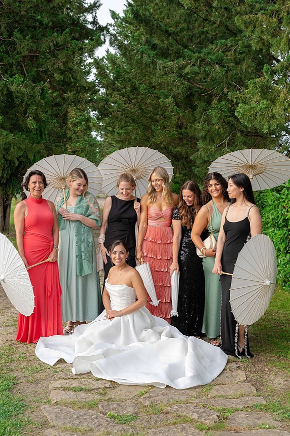 Bridal party portrait with bride and bridesmaids holding paper parasols, posed on a stone path beside green hedges and trees