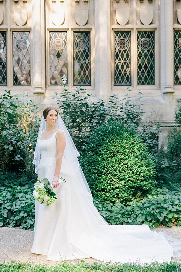 Bridal portrait of a bride in a white gown with wedding veil, holding a greenery bouquet on a walkway by a stone facade with leaded windows