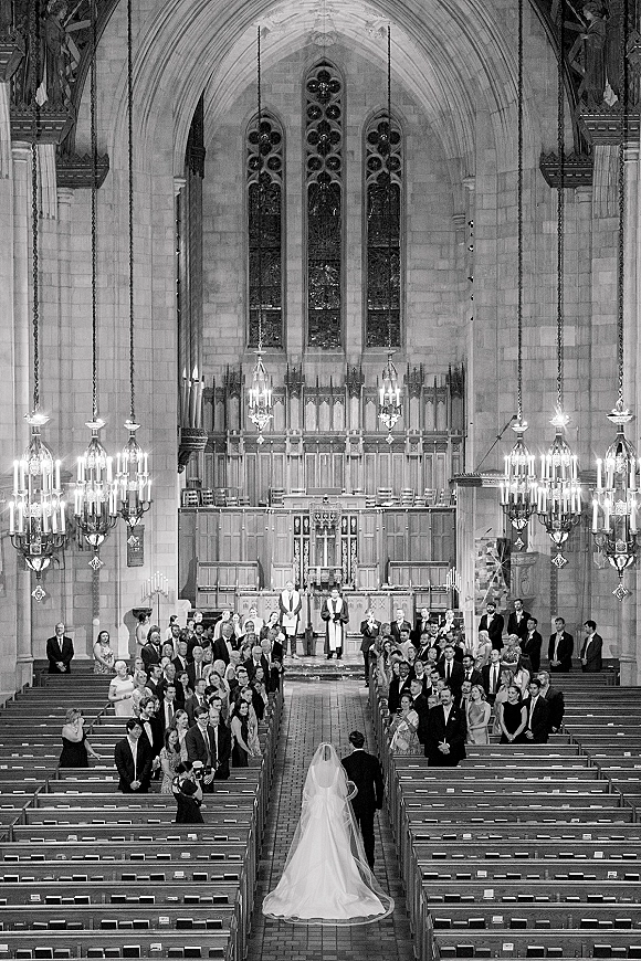 Wedding ceremony in a cathedral as the bride walks down the aisle in a long veil toward the groom waiting at the candlelit altar