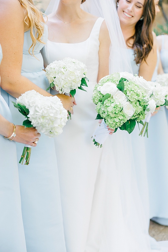 Bridesmaid bouquets of white hydrangeas and roses held by women in light blue dresses beside a veiled bride outdoors with blurred foliage