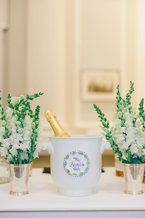Champagne bucket at a wedding champagne station with a bottle in ice, monogram sticker, and white floral arrangements in silver pots on a table