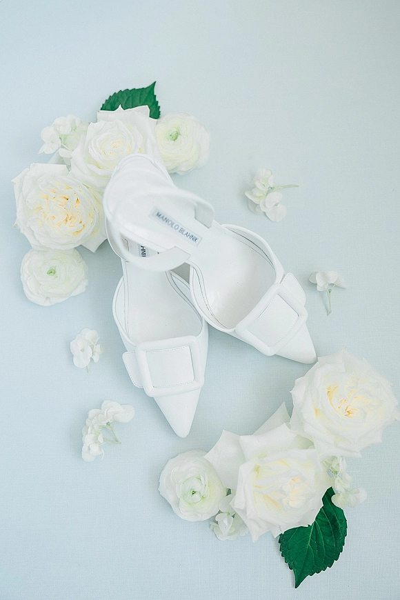 Bridal shoes in a wedding shoe flat lay, white pointed-toe heels with ankle straps and buckle detail surrounded by white roses on light blue surface
