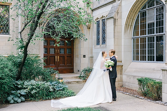 Couple portrait of bride and groom embrace, her long veil and gown train flowing as they hold a bouquet by a stone building entrance.
