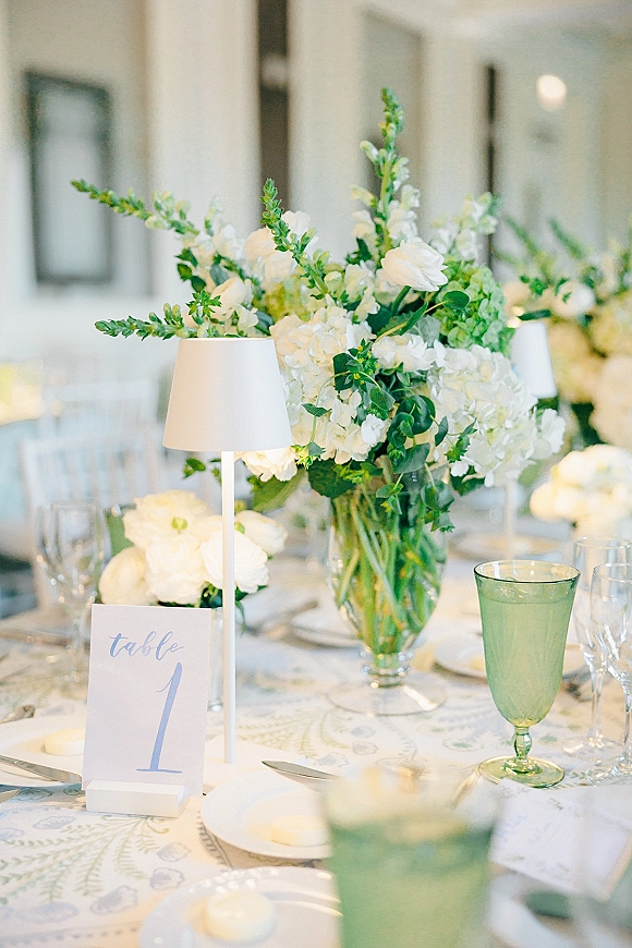 Reception tablescape with a wedding table centerpiece of white hydrangeas and greenery in a clear vase, with green goblets and table number card