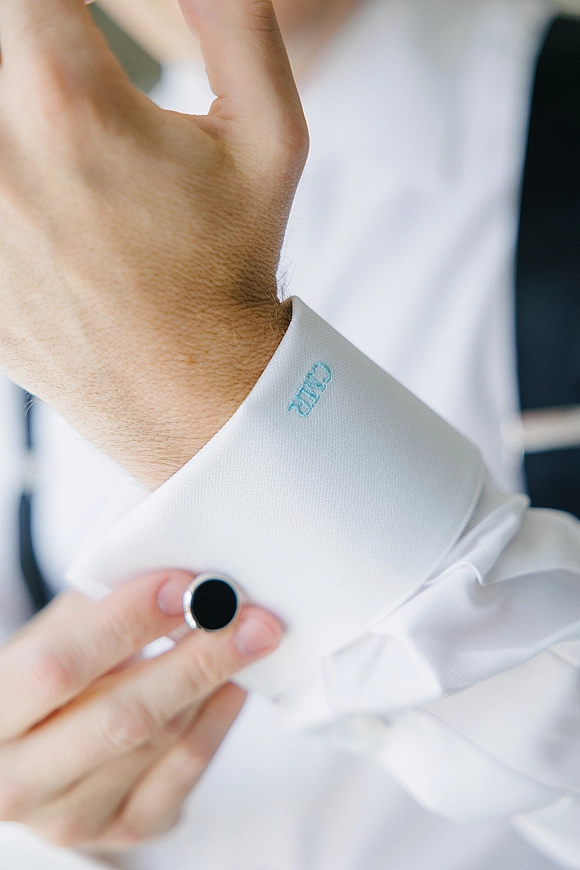 Groom cufflinks close up on a white French cuff dress shirt with monogram embroidery, blurred suit jacket in soft indoor light