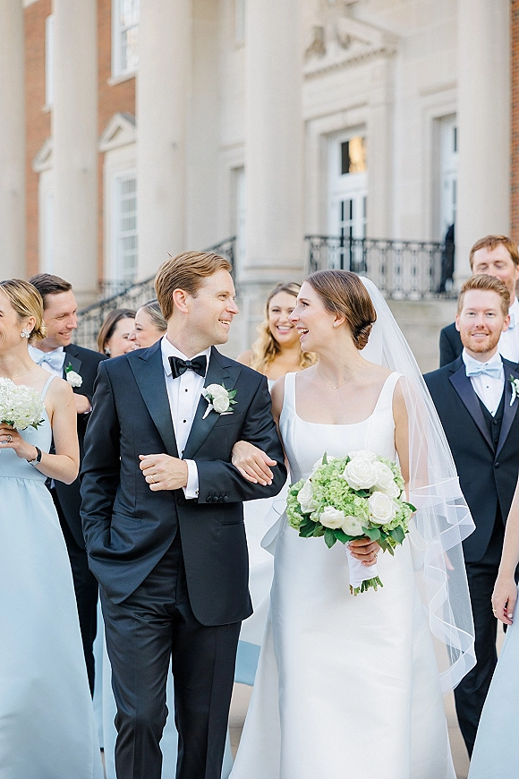 Wedding party portrait of bride and groom with wedding party walking on stone steps, bride holding bouquet with a long veil by columns