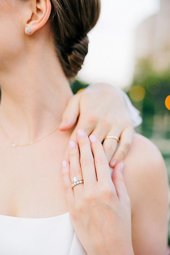 Wedding rings close-up on bride’s hand, engagement ring sparkling beside band as she rests fingers on shoulder, greenery bokeh behind
