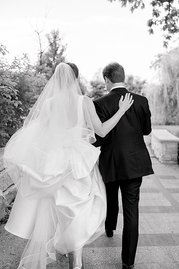 Couple portrait of bride and groom walking away on a stone garden walkway, her long veil trailing over a ball gown beside trees