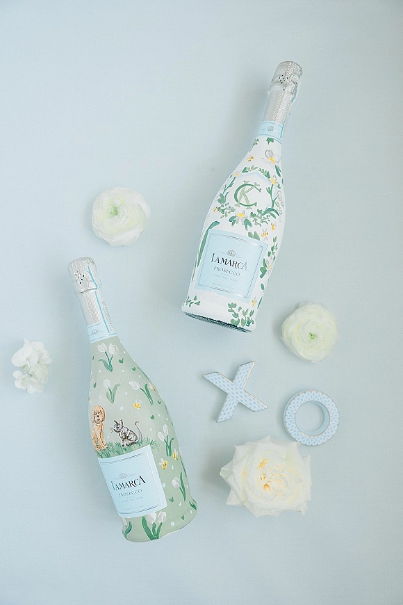 Prosecco bottles with wedding prosecco bottles labels, white flowers, and X and O decor arranged on a light blue surface flat lay