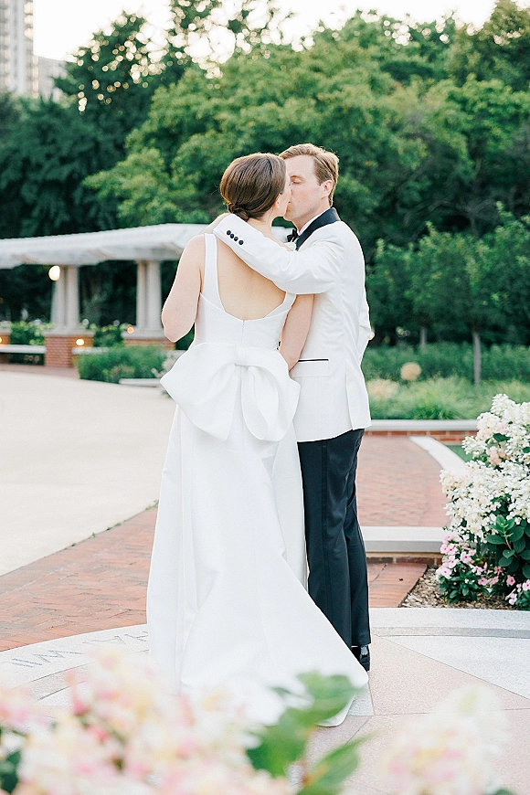 Wedding kiss portrait of bride in open-back gown with bow detail and groom in white tuxedo, embracing on a brick garden walkway near columns