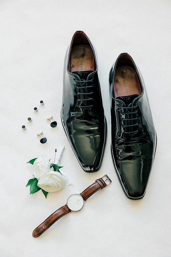 Groom accessories flat lay with black patent dress shoes, cufflinks, wristwatch, and boutonniere arranged on a white surface