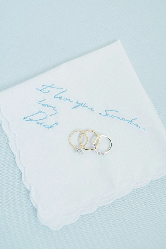 Wedding rings in a wedding ring flat lay, gold bands with a diamond ring on a white napkin with blue embroidery over light blue linen