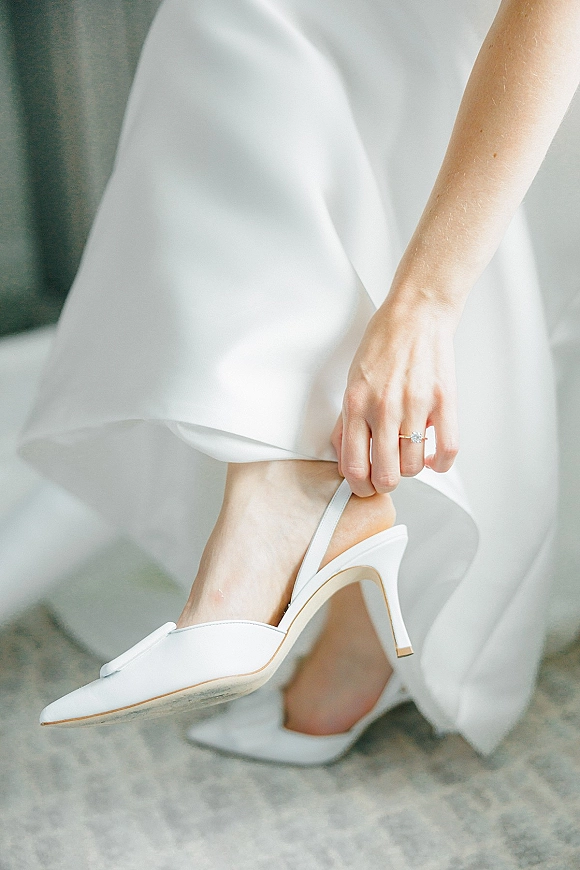 Bridal shoes, white wedding heels on a stone floor as the bride adjusts slingback straps, engagement ring glinting by her gown hem