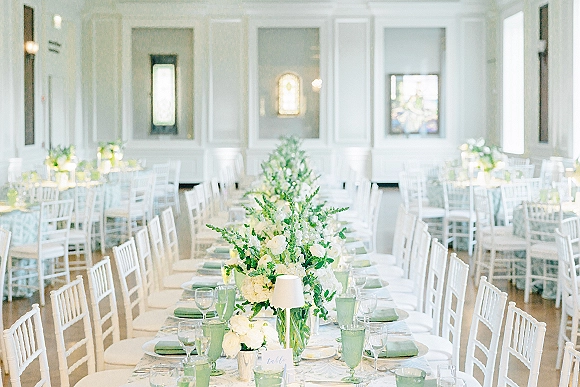 Reception tablescape with a wedding long table setup featuring white floral and greenery centerpiece, green goblets, sage napkins, candles, and lamps in a paneled room