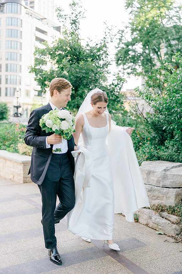 Couple portrait of bride and groom walking, her veil trailing as he carries a white bouquet beside a stone wall and trees