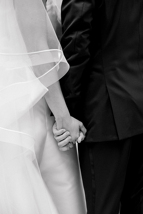 Couple holding hands in a wedding hand holding photo, bride’s veil and gown beside groom’s suit against a neutral backdrop