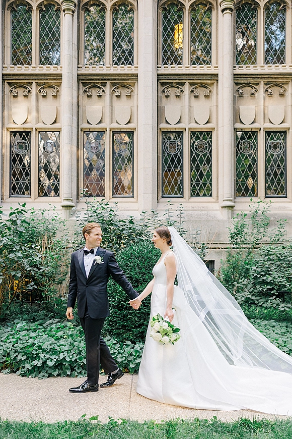 Couple portrait of bride and groom holding hands, her long veil trailing as they walk past a stone building with gothic windows