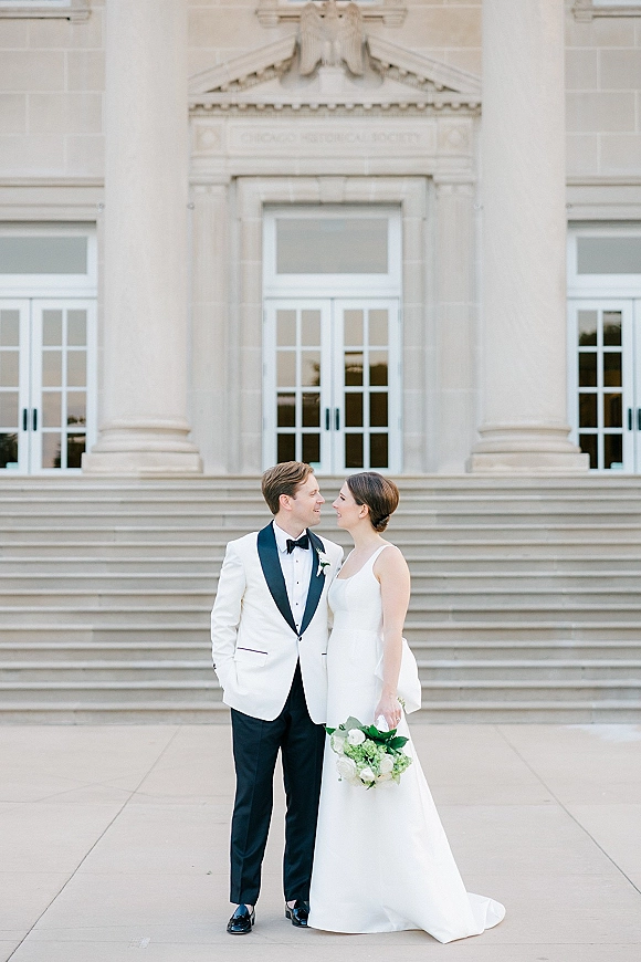 Couple portrait of bride and groom in black tie, holding a white and green bouquet on stone steps before a grand columned facade