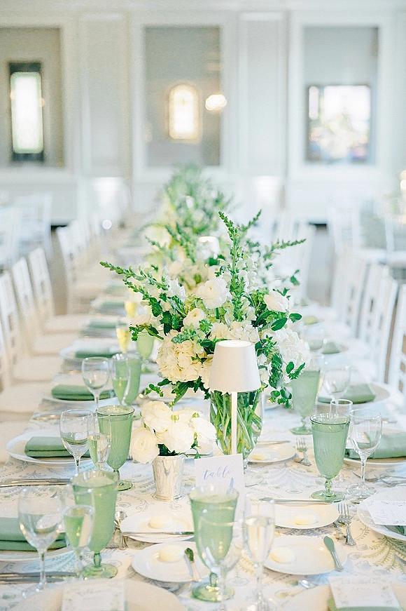 Reception tablescape with wedding table centerpiece of white blooms and greenery, green goblets, candles and place cards in a bright room