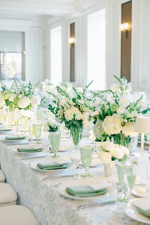 Reception tablescape with white and green tablescape details, white floral centerpieces in clear vases, green goblets, and mini lamps in a bright room