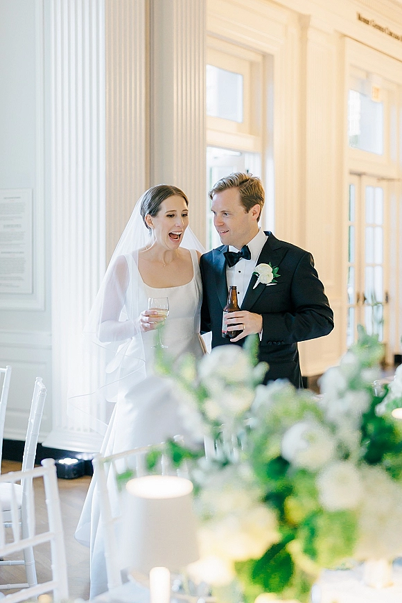 Wedding couple portrait of bride and groom reception, laughing with wine glass and beer bottle at a bright room with white columns