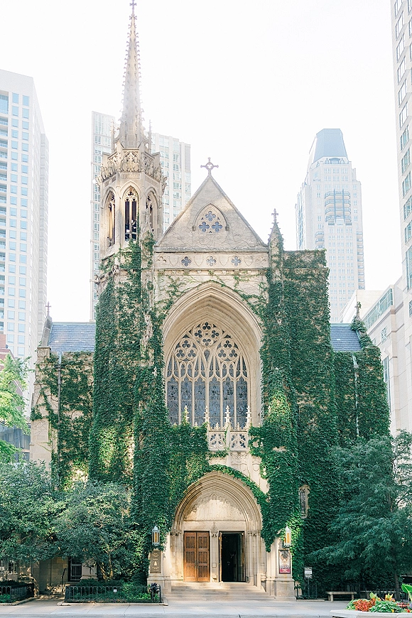 Church exterior with ivy climbing a stone gothic facade, arched window and wooden doors, set along a city street near skyscrapers and trees