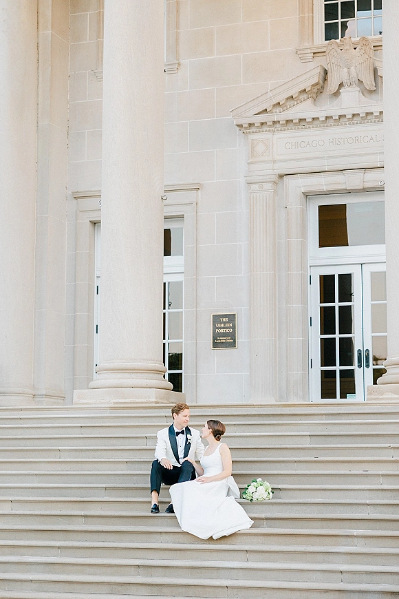 Couple portrait of bride and groom sitting on steps, gazing at each other in black tie with bouquet before grand columns and doors