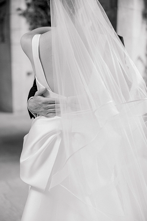 Wedding kiss portrait with a veil over couple kiss as the groom holds the bride’s waist on a blurred outdoor walkway by a wall