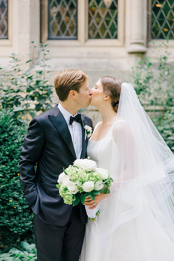 Wedding kiss portrait of bride and groom kissing, her long veil and white rose bouquet against a stone building with leaded windows