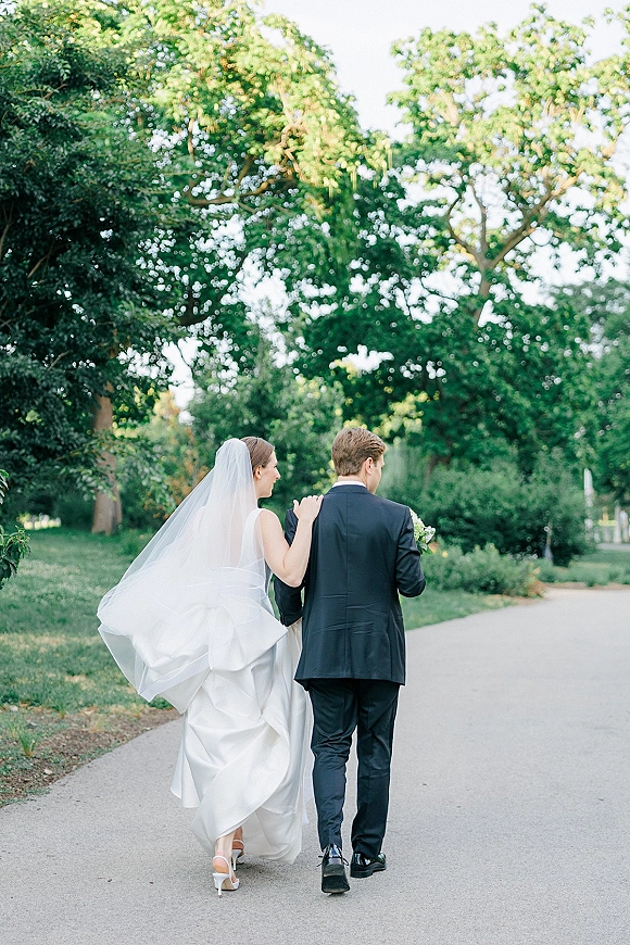 Couple portrait of bride and groom walking away on a tree-lined park path, long veil blowing as he carries a white bouquet
