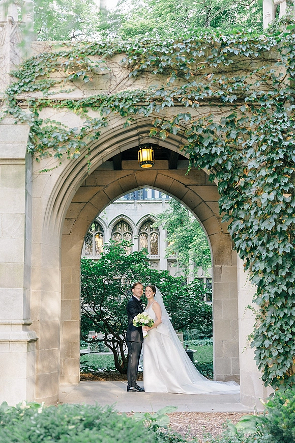 Couple portrait of bride and groom portrait under an ivy-covered stone archway, bride in veil holding bouquet beside groom in tuxedo