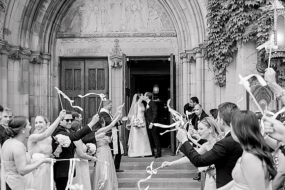 Wedding recessional as bride and groom kiss at a church exit, bouquet and veil flowing while guests wave ribbon wands on stone steps