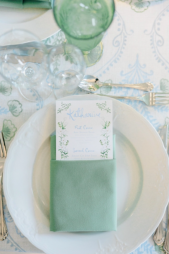 Reception tablescape with a wedding place setting featuring a place card on stacked plates, menu on folded napkin, green goblet on patterned tablecloth