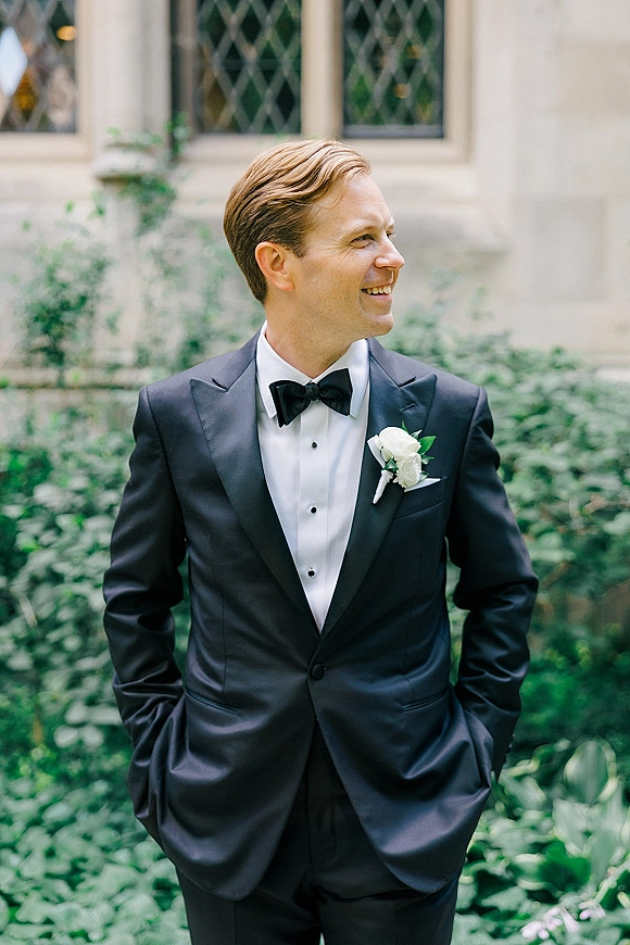 Groom portrait in a black tuxedo with bow tie and white boutonniere, hands in pockets, looking away by garden greenery and stone facade