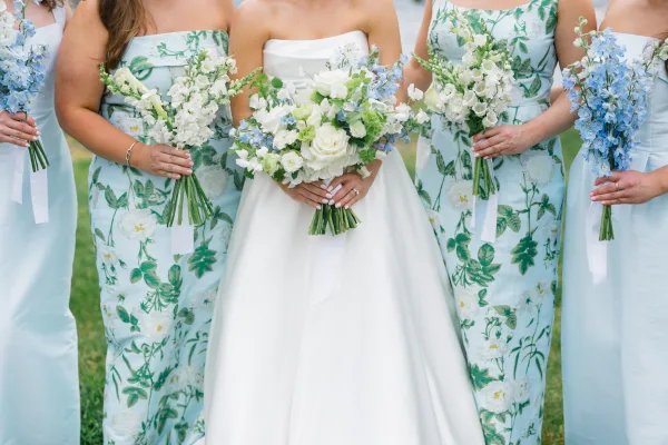 Bridesmaid dresses line up in coordinated looks, including blue bridesmaid dresses, holding bouquets on a grass lawn beside the water.