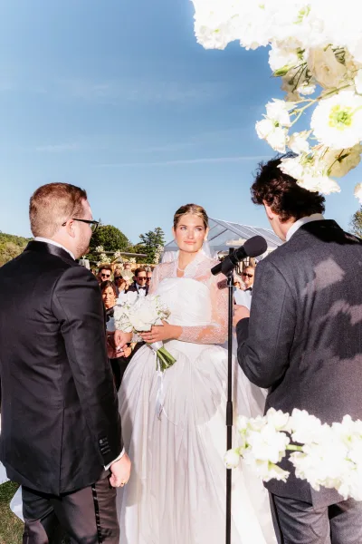Wedding vows as bride and groom hold hands at an outdoor wedding ceremony under a floral arch, bride with veil and white bouquet by microphone