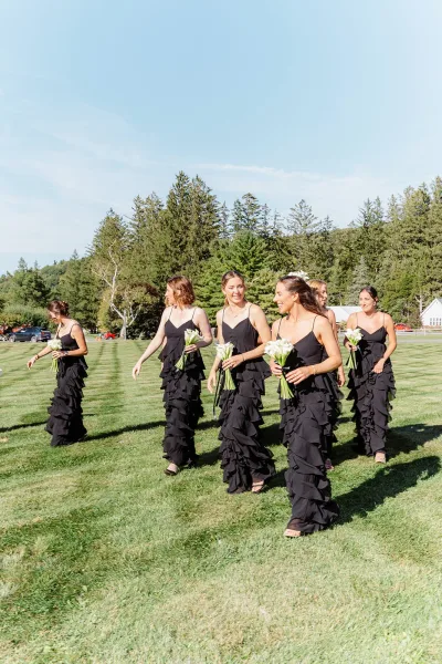 Bridesmaid procession in black dresses holding white calla lily bouquets, walking across a grass lawn with trees and blue sky behind