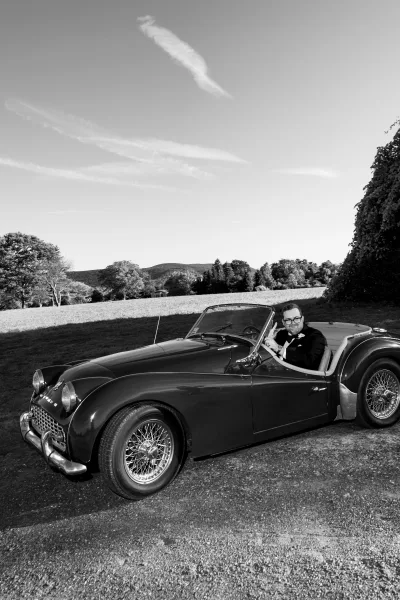Groom portrait in a vintage car, wearing a black tuxedo with bow tie and boutonniere beside an ivy-covered wall under cloudy sky