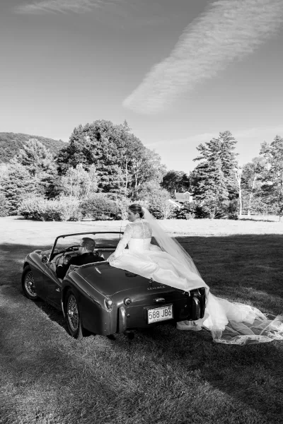 Wedding couple portrait with bride in vintage car, lace sleeves and long veil flowing as groom sits by a countryside field with mountains