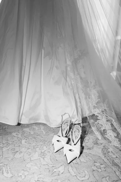 Wedding shoes with white bow slingback heels tucked under a bridal gown hem, lace veil draped over them on patterned carpet indoors