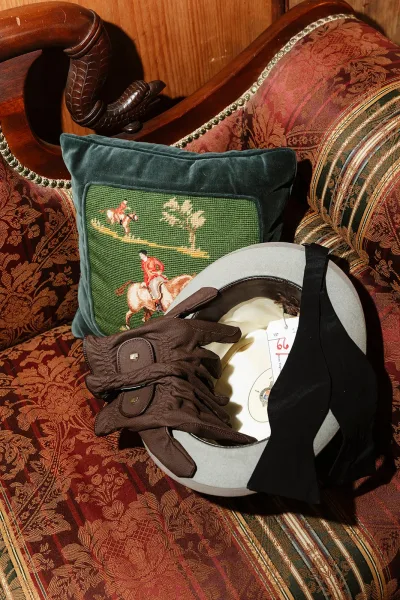 Groom accessories with a wedding groom hat and brown leather gloves arranged on an upholstered sofa with an embroidered pillow and wood paneling backdrop