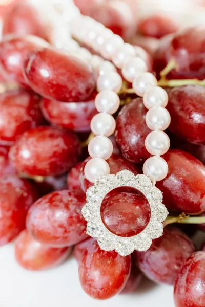 Wedding rings with a diamond halo ring beside a pearl bracelet and red grapes on a white surface, styled as a bridal detail flat lay