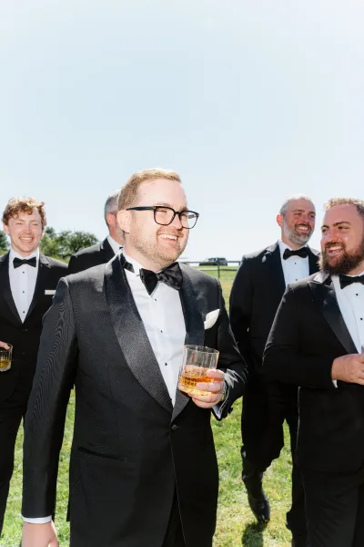 Groom and groomsmen in black tuxedos raise whiskey glasses for a toast on a sunny lawn with trees and blue sky behind them