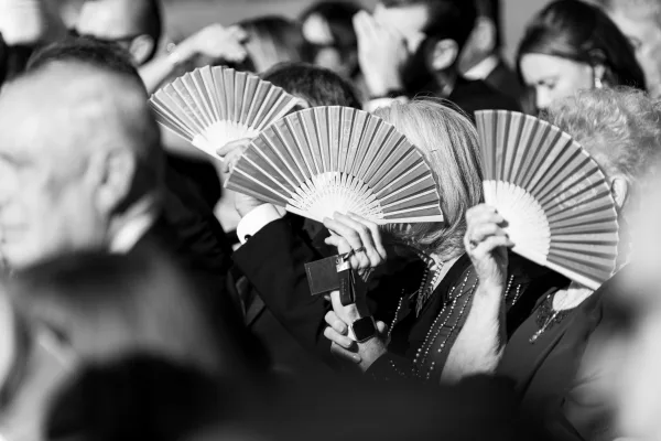 Wedding guests reacting during the ceremony, smiling in indoor seating while holding hand fans amid a lively crowd