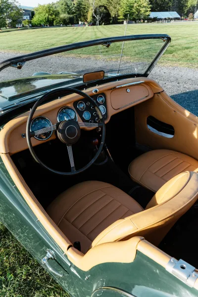 Vintage wedding car interior with tan leather seats and steering wheel, parked by a grassy field and white fence under trees