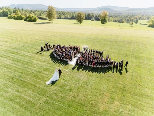 Outdoor wedding ceremony aerial view shows semi-circle seating around a white aisle runner and floral arch on a mountain lawn