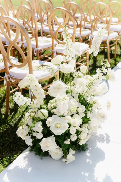 Ceremony aisle decor with a white aisle runner on grass, lined with cross back chairs and low white rose and delphinium florals in sunlight
